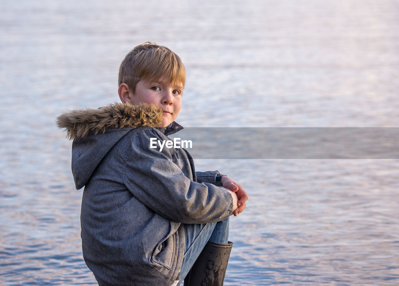 Portrait of boy wearing warm clothing sitting on rock by lake during sunset