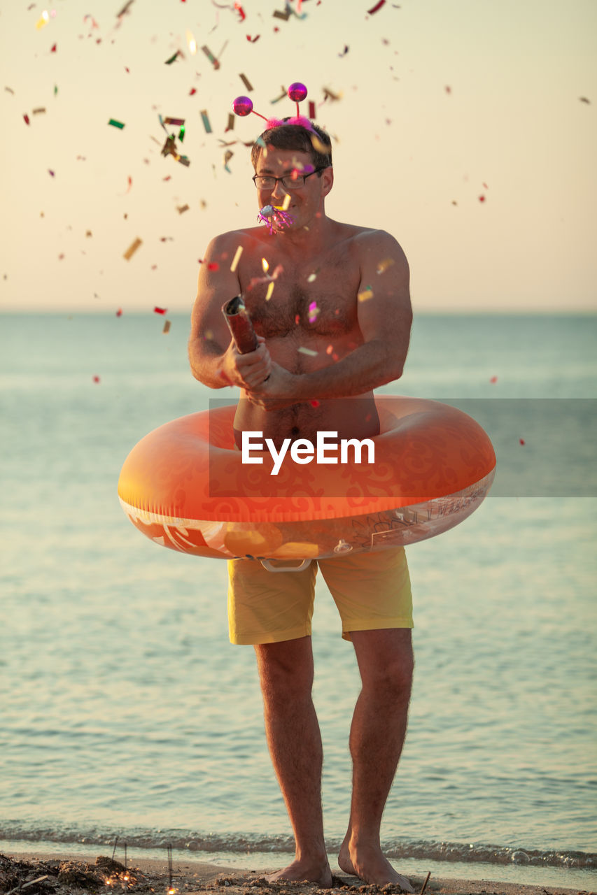 Full length of shirtless man with inflatable ring and confetti standing at beach during sunset