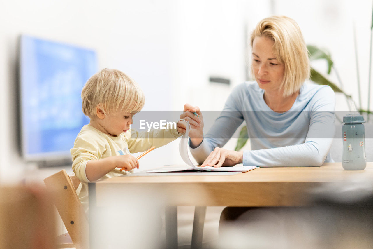 child, childhood, women, blond hair, female, adult, men, two people, family, table, indoors, sitting, parent, togetherness, person, learning, one parent, casual clothing, conversation, smiling, baby, domestic life, bonding, lifestyles, toddler, happiness, emotion, writing, education, home interior, selective focus, homework, positive emotion, communication, furniture, domestic room, care, day, looking
