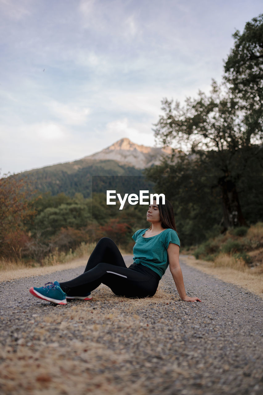 Full body side view of relaxed young female traveler with eyes closed sitting on road and enjoying freedom during adventure in mountainous countryside of spain