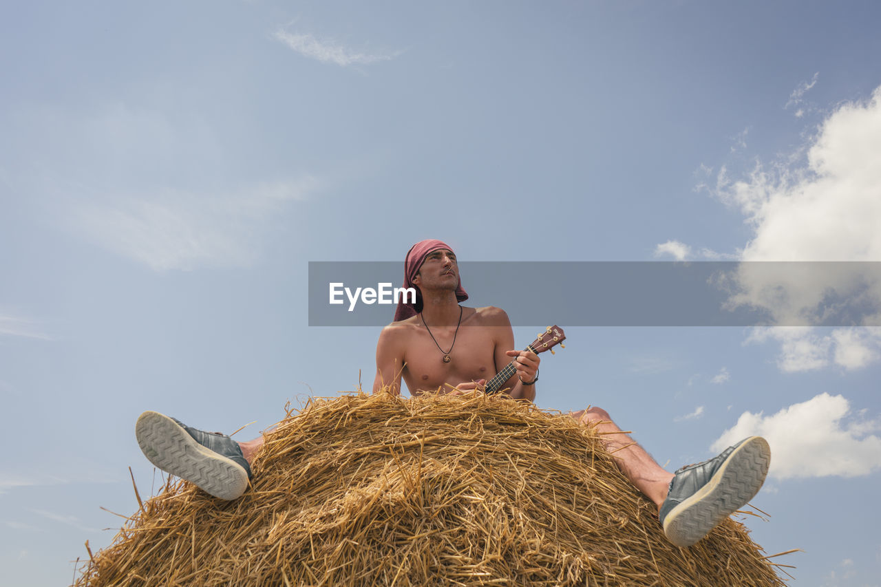 Low angle view of shirtless man playing ukulele on hay bale against blue sky