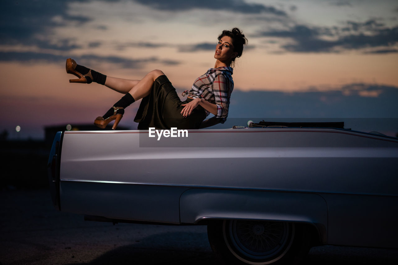 Portrait of woman standing by car against sky during sunset