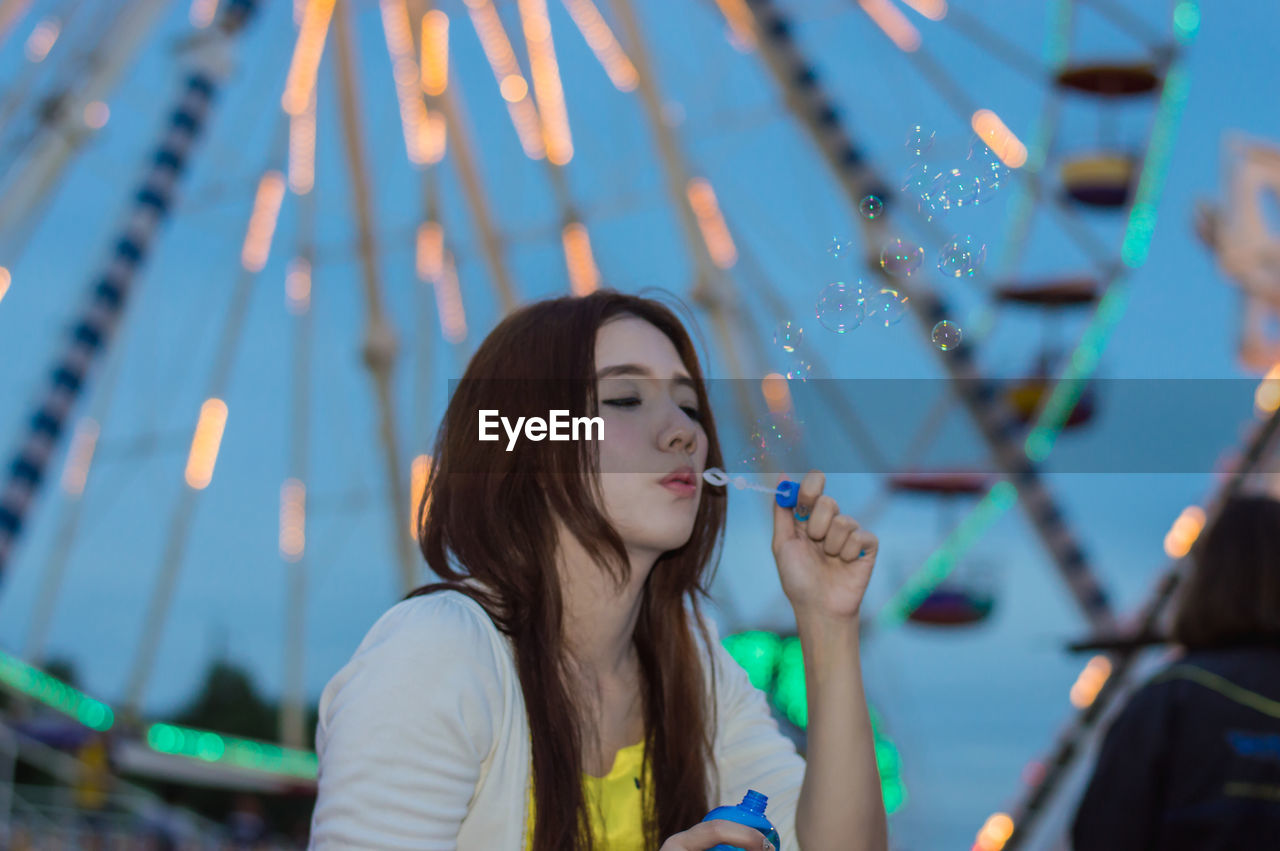 Young woman blowing bubbles standing against ferris wheel in amusement park