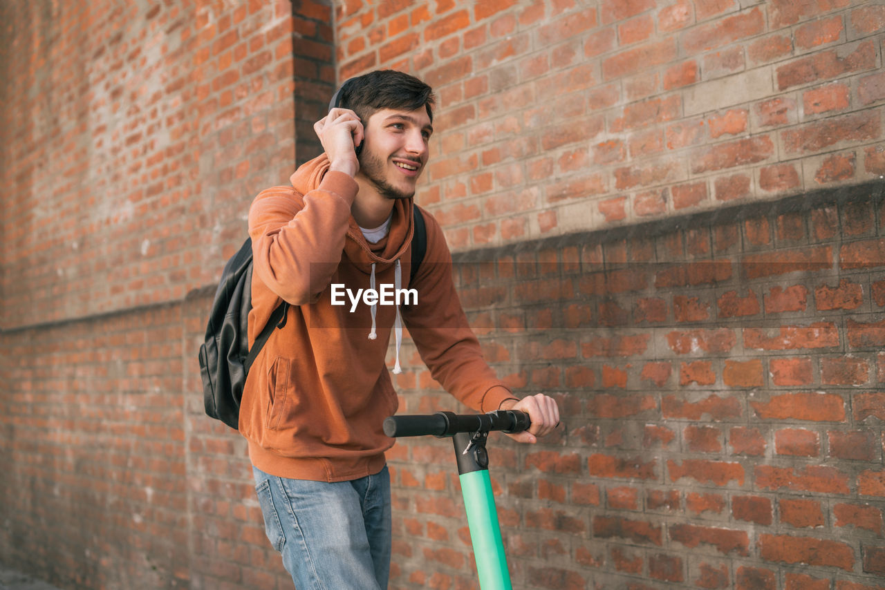 Young man listening music while riding push scooter against brick wall