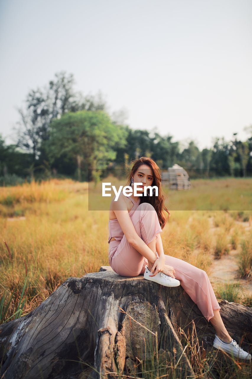 Portrait of young woman sitting on tree stump against clear sky