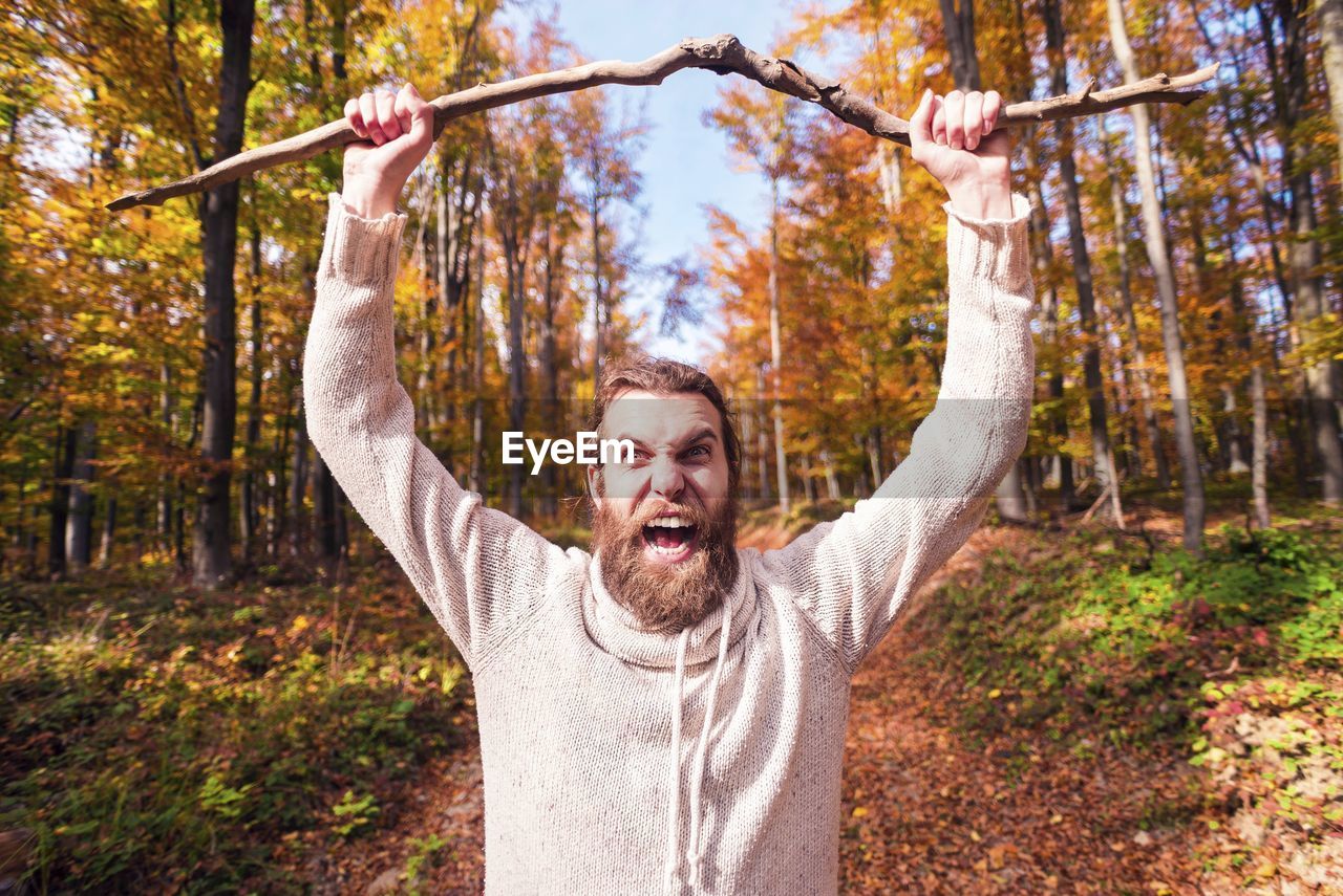 Portrait of bearded man holding stick screaming while standing against trees in forest