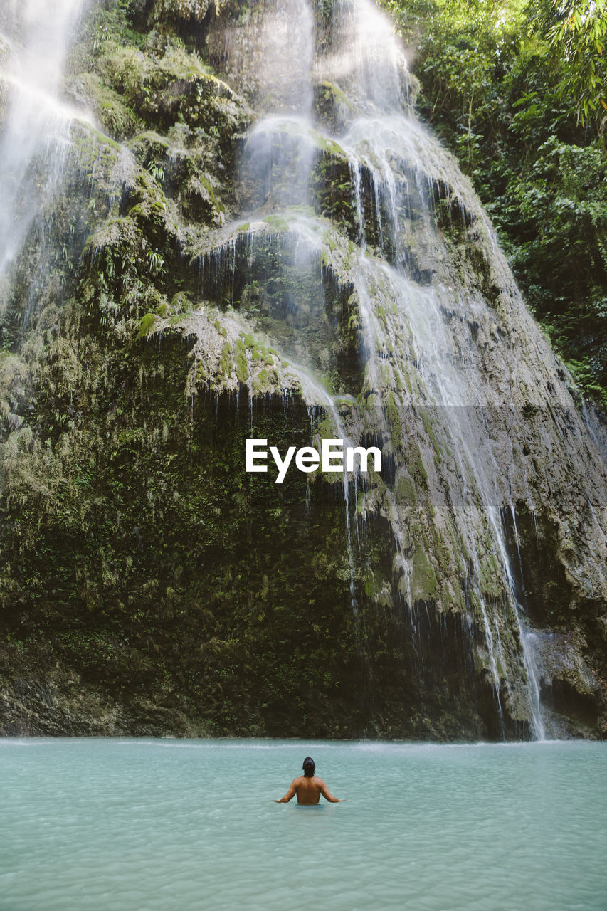 Rear view of shirtless man swimming in river by waterfall