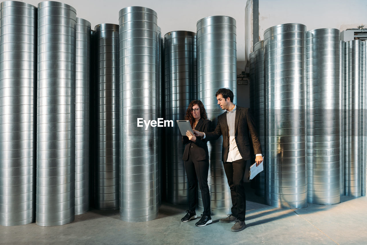 Busy male and female colleagues in formal clothes using tablet together while working in storehouse with metal barrels