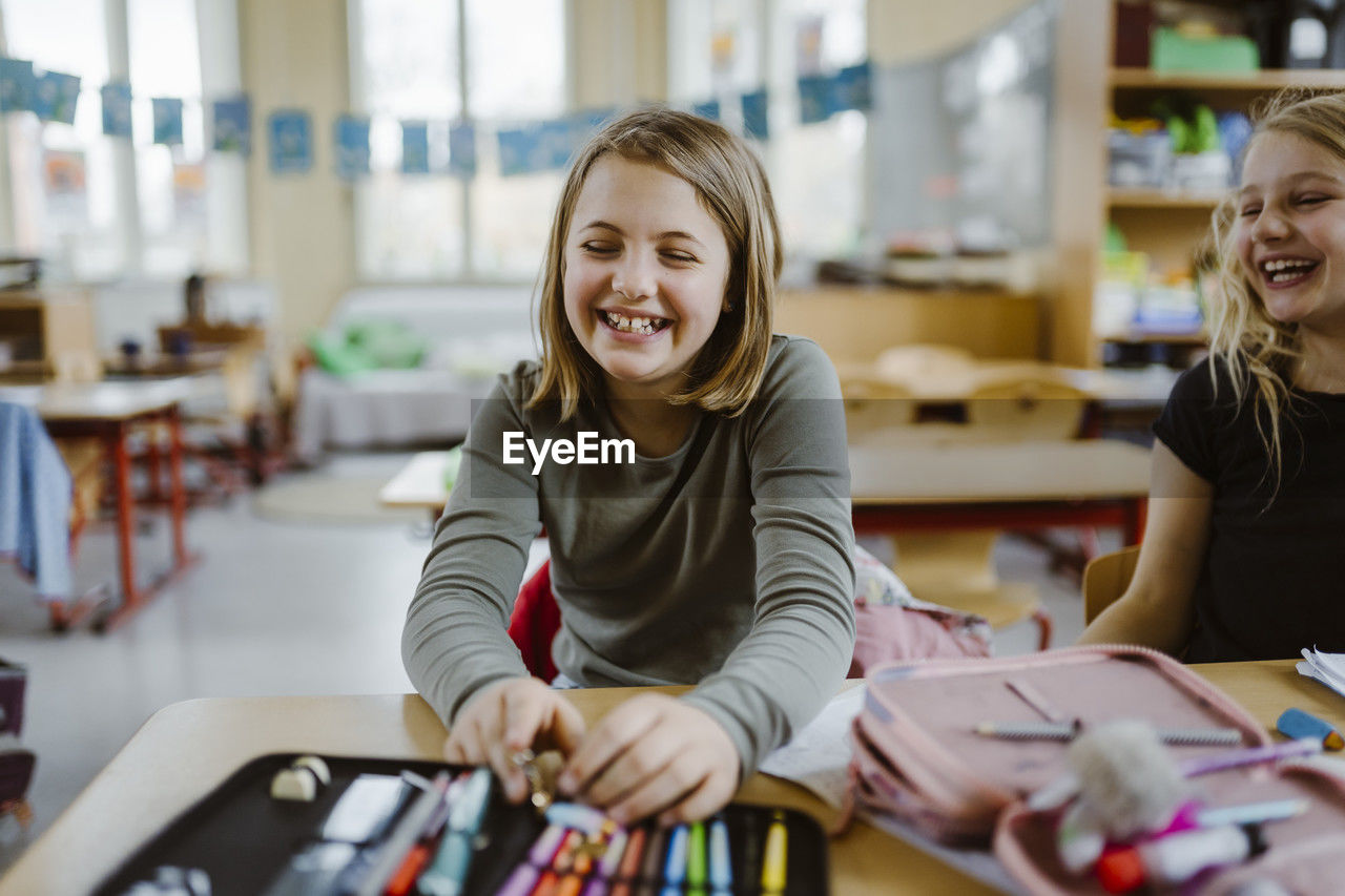 Happy schoolgirl laughing while sitting with friend at desk in classroom