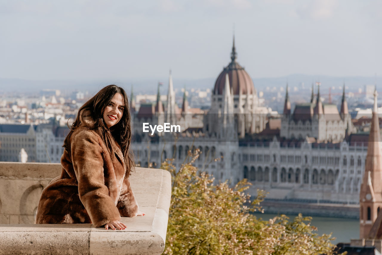 Portrait of beautiful young woman on balcony overlooking hungarian parliament in budapest, hungary