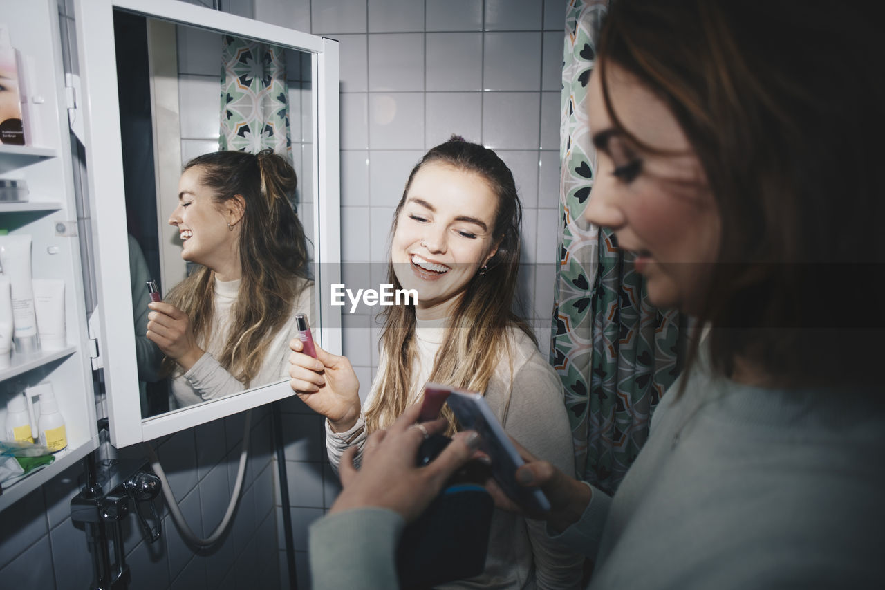 Happy female friends with mobile phone and lip gloss at dorm bathroom