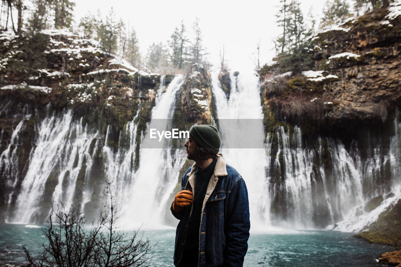 Man standing on picturesque scenery of powerful waterfall with pool flowing among snowy forest in mountainous terrain in winter day in usa looking away