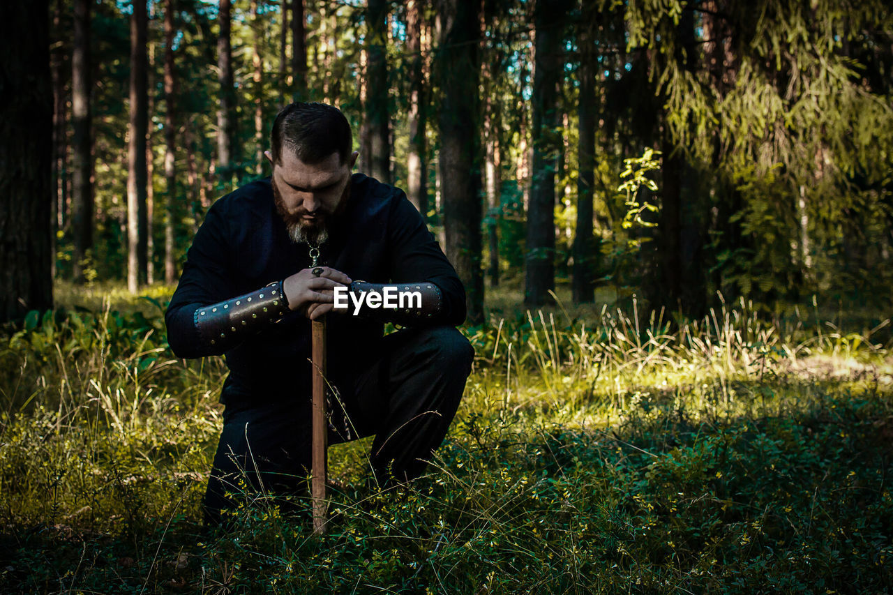 Man with axe crouching on field against trees