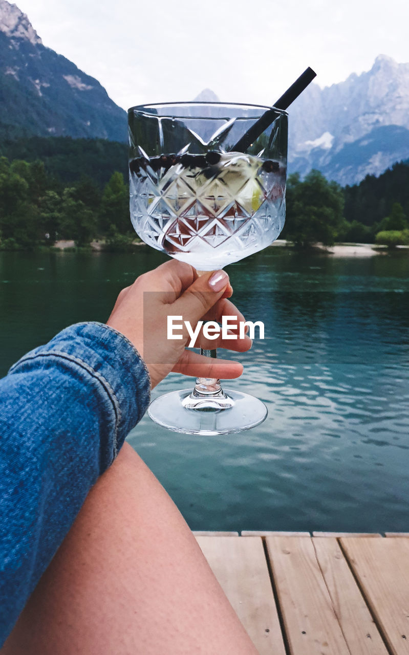 Personal perspective of woman holding glass with drink. lake, mountains in background.