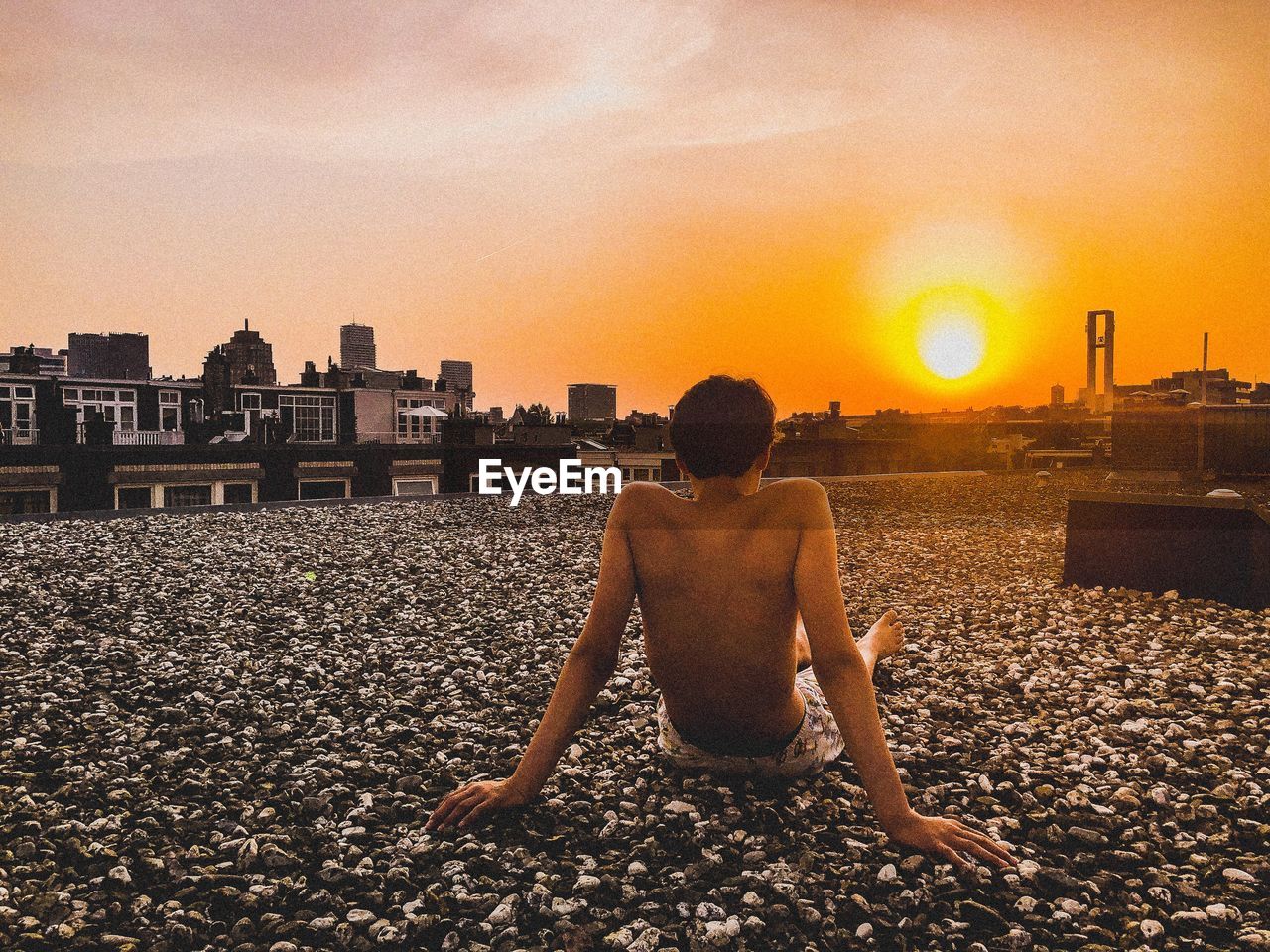 Rear view of shirtless boy sitting on stones during sunset