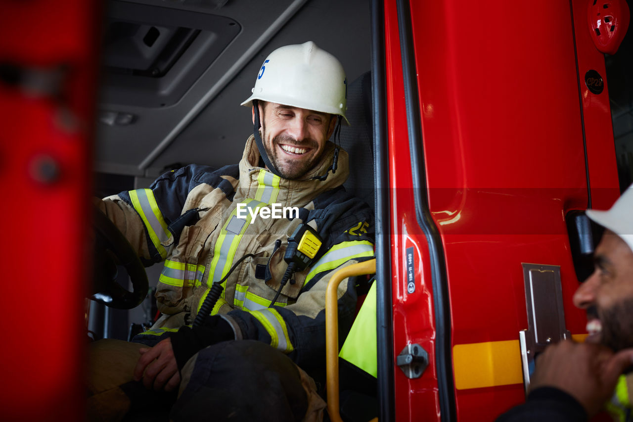 Smiling firefighter sitting in fire engine while talking to coworker