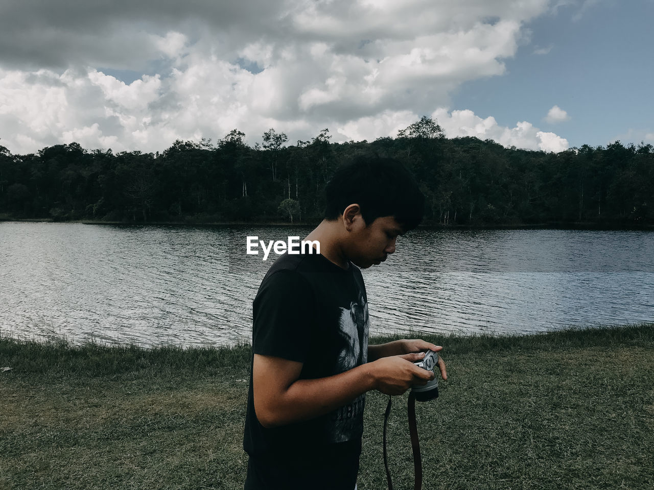 Side view of man holding camera standing by lake against sky
