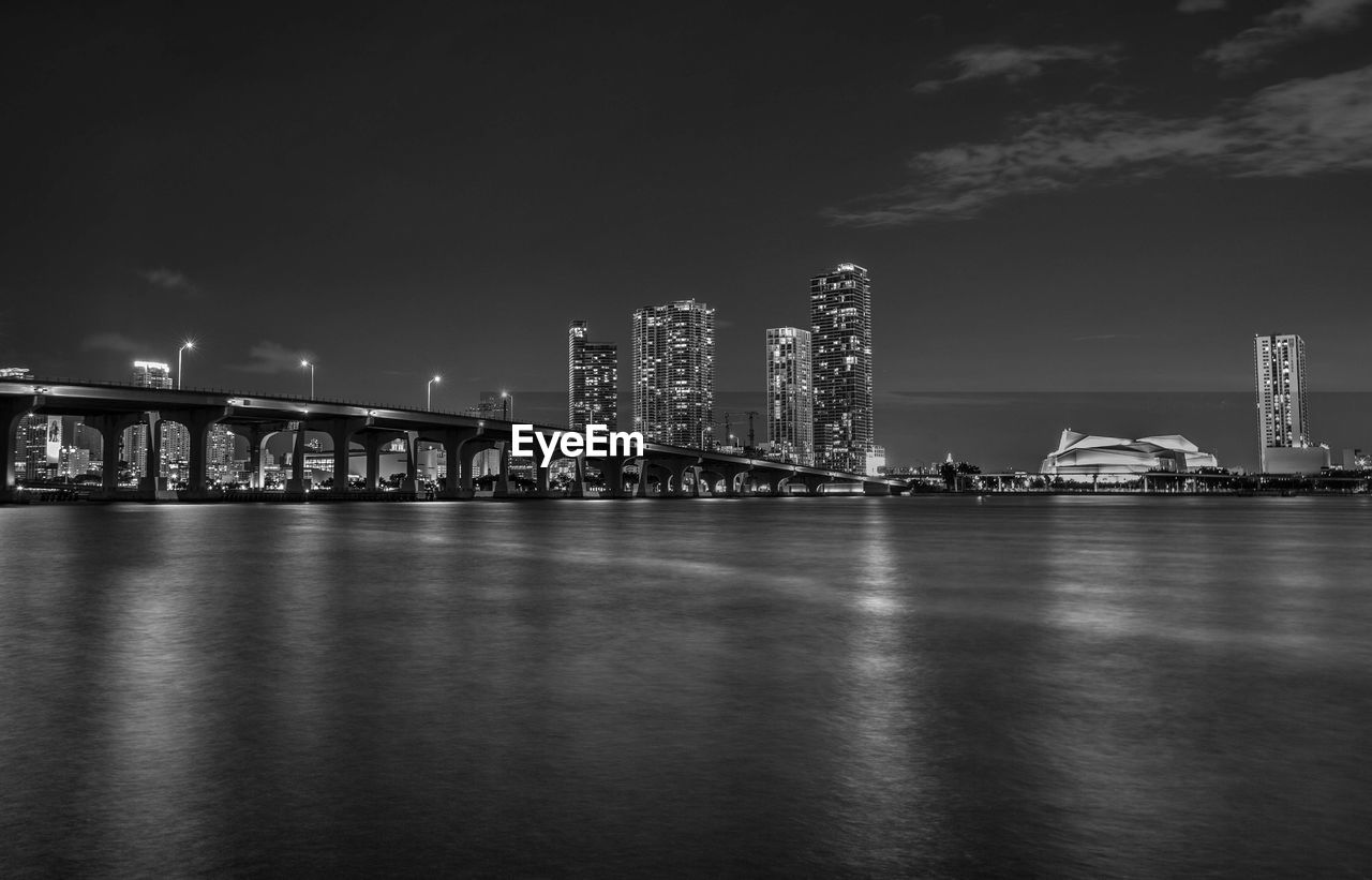 Illuminated bridge over river by buildings against sky at night