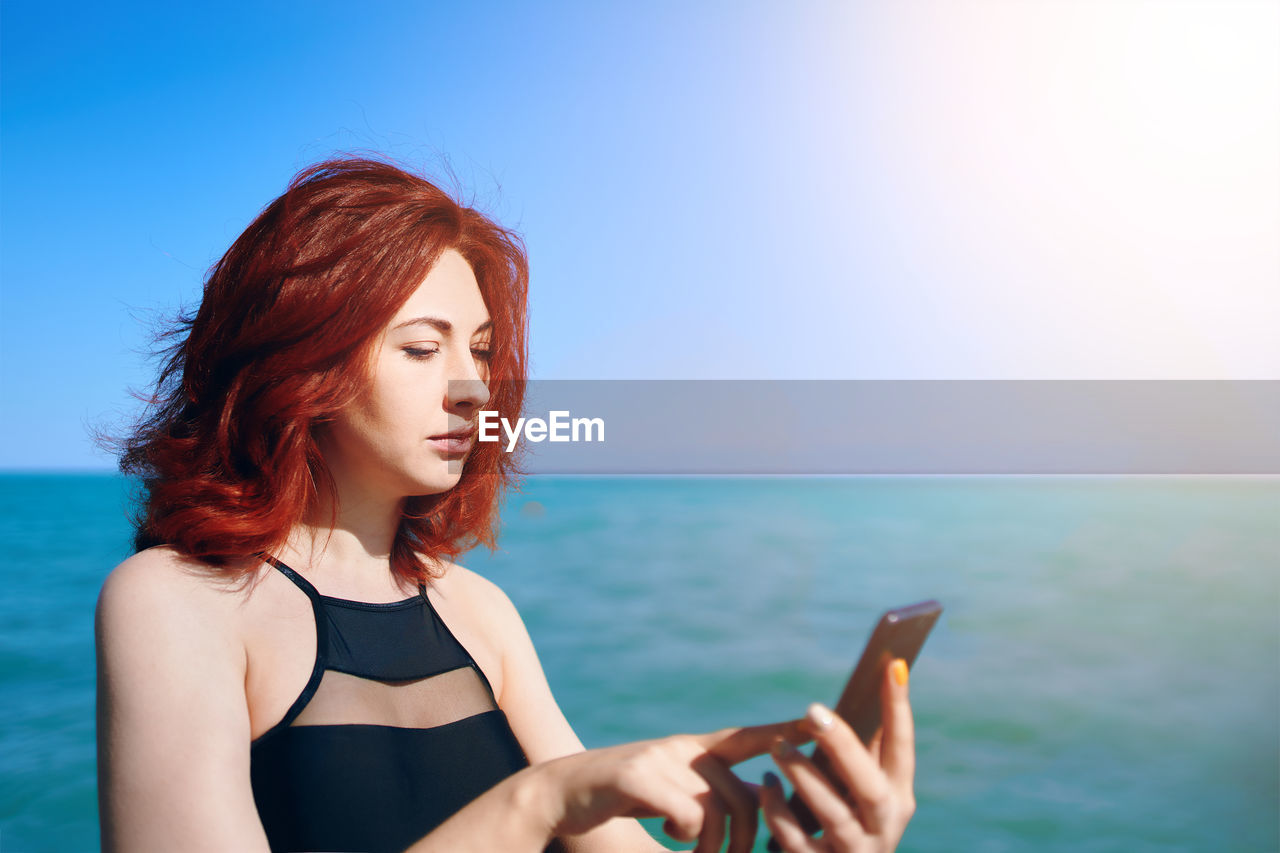Beautiful young woman using smart phone against sea