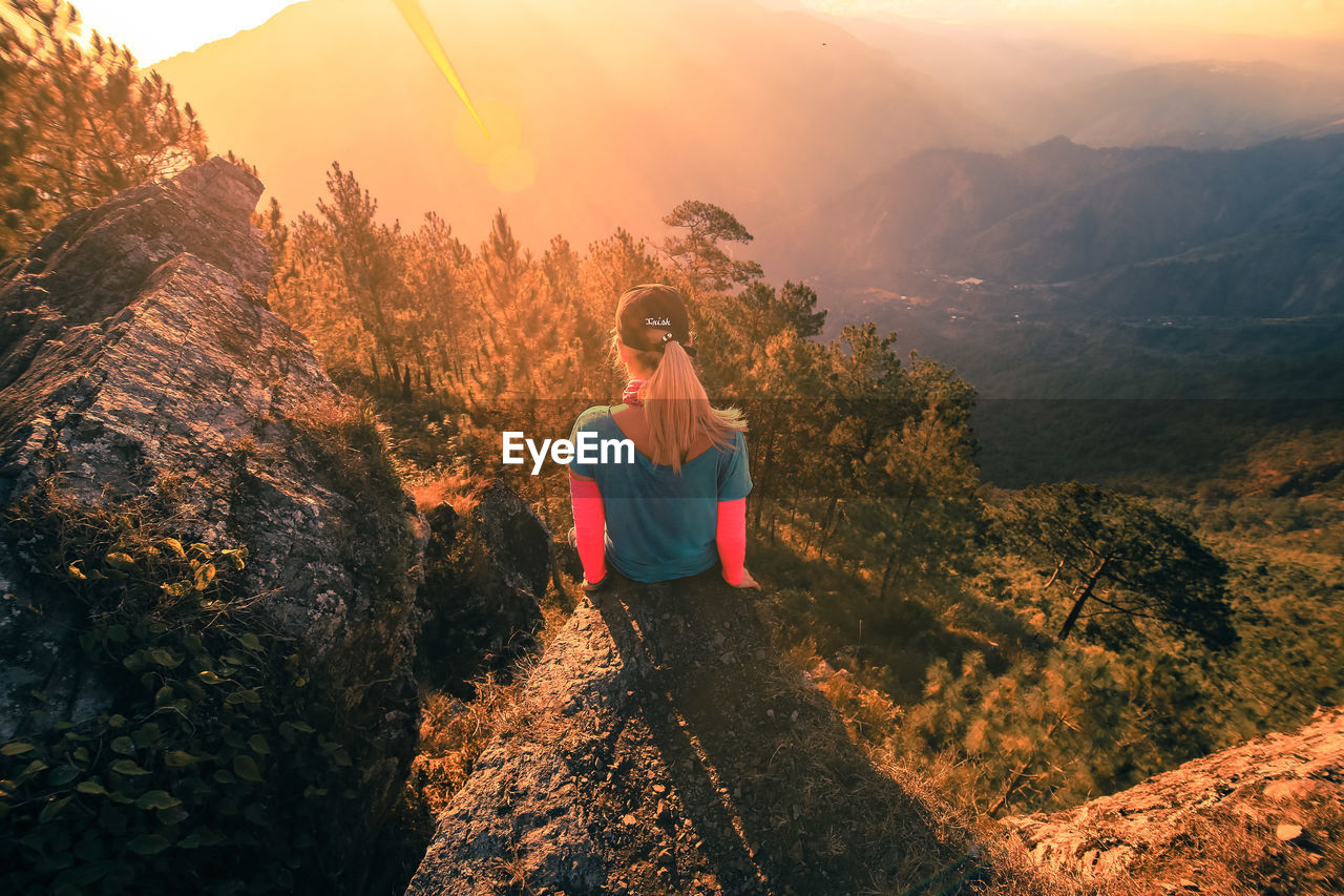 Rear view of teenager sitting on cliff against mountains