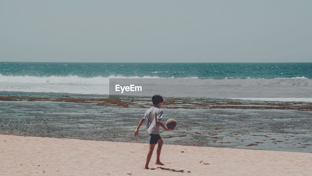 Boy playing with ball at beach against clear sky