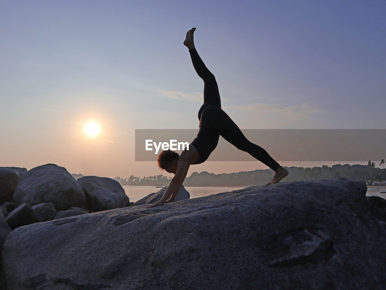 Woman practicing yoga, doing eka pose on adho mukha svanasana, on rock by beach at sunrise