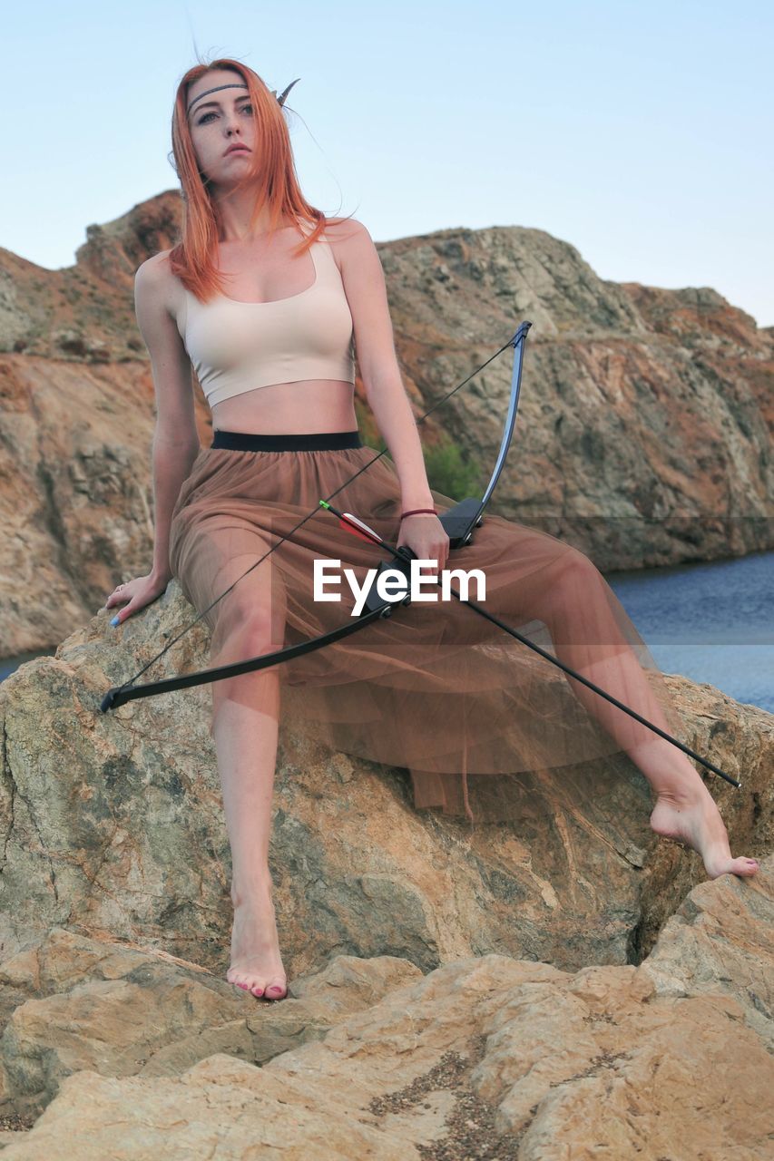Full length of young woman holding bow and arrow while sitting on rock