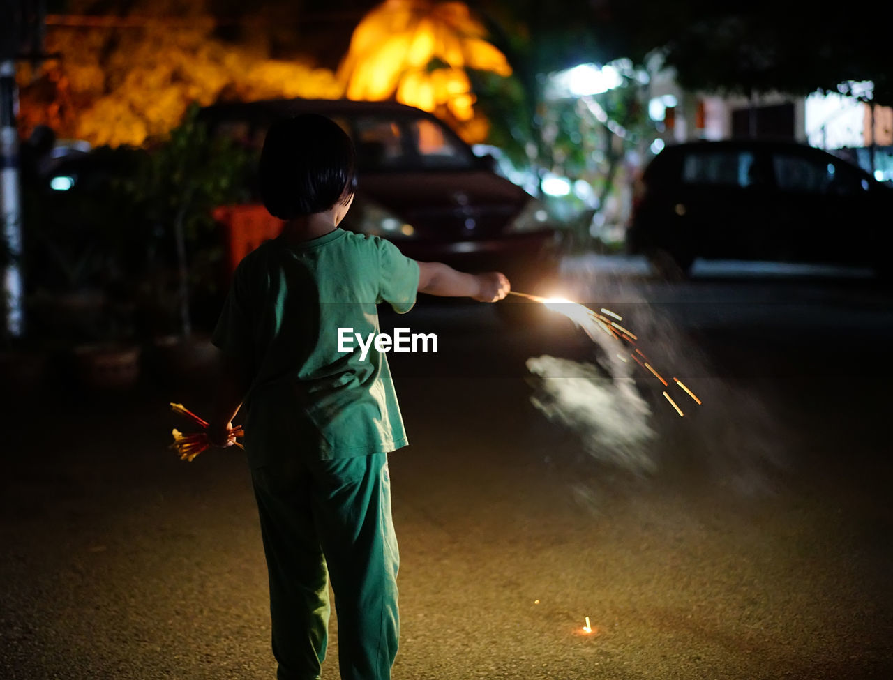 Rear view of girl holding illuminated firework on street in city at night