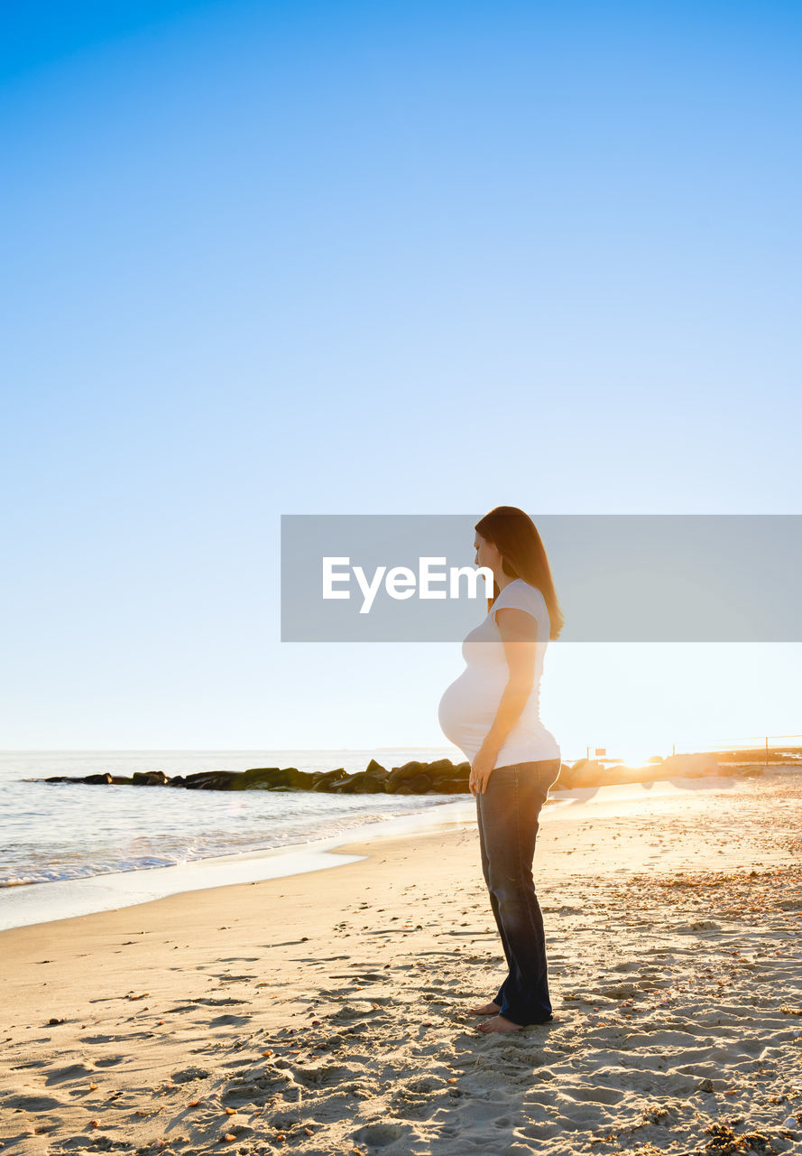 Full length of young pregnant woman 3rd trimester standing at beach against sunset clear sky