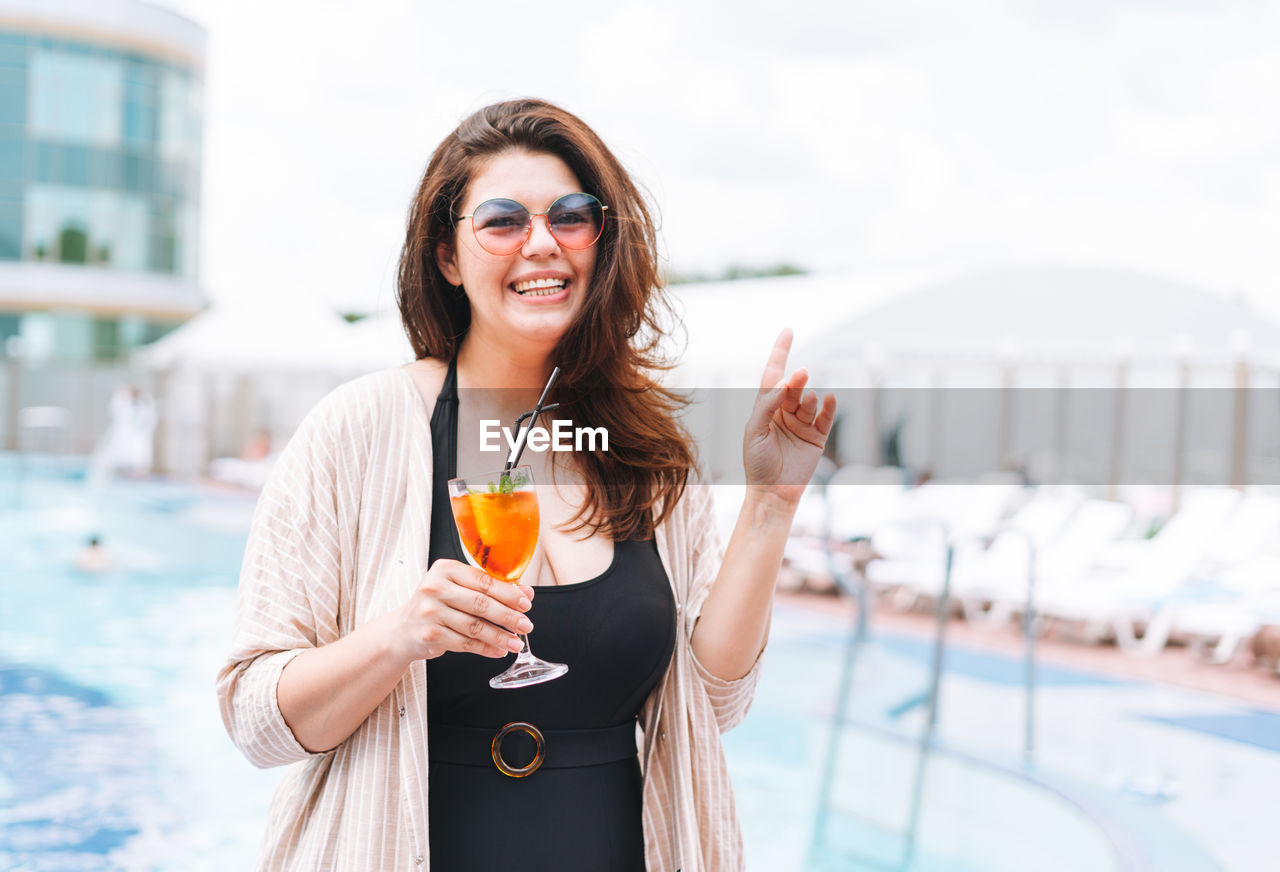 Stylish woman plus size body positive in sunglasses drinking with glass of cocktail near pool