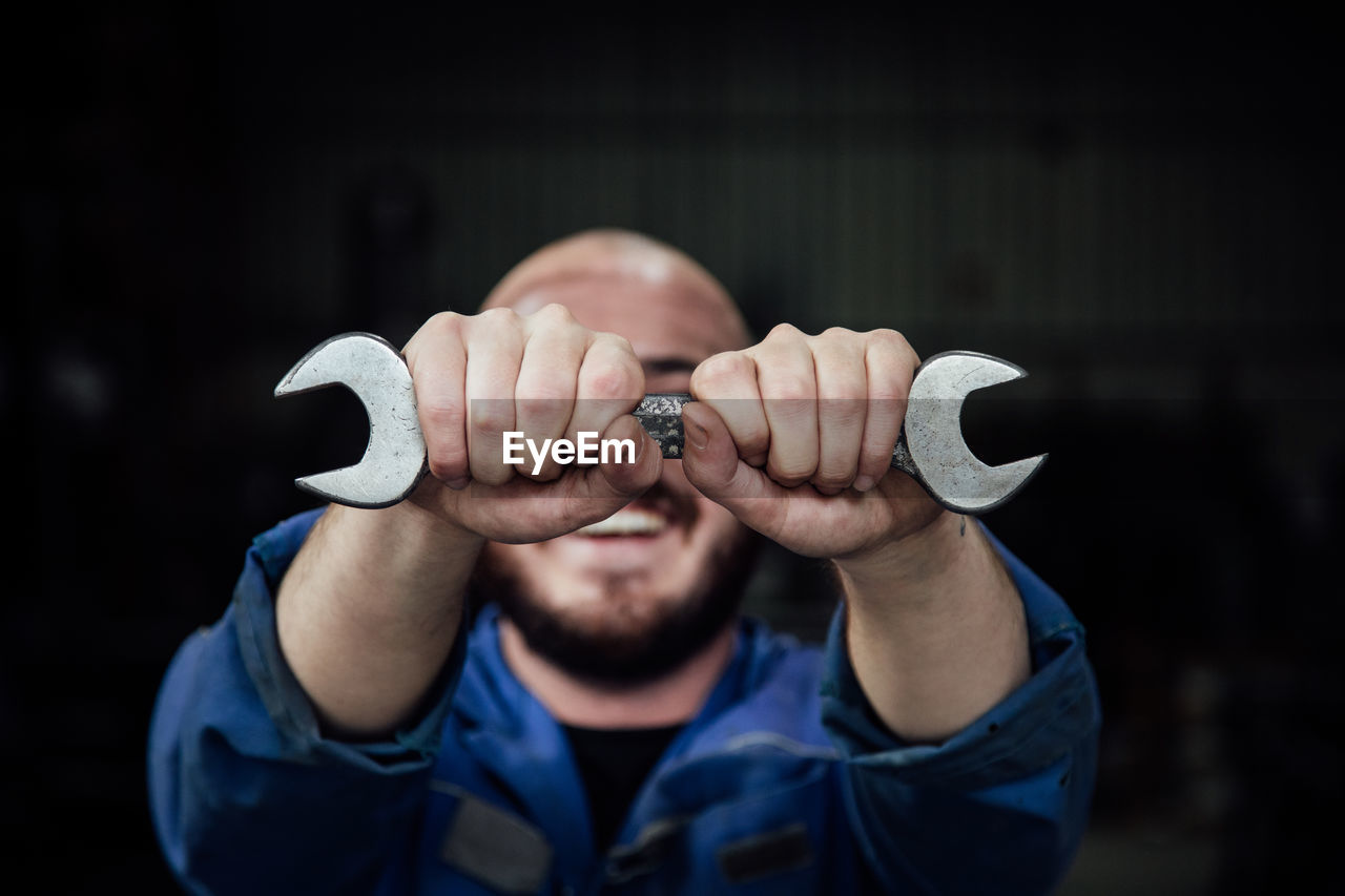 Faceless male mechanic in blue overall covering face with metal spanner in hands looking at camera on black background