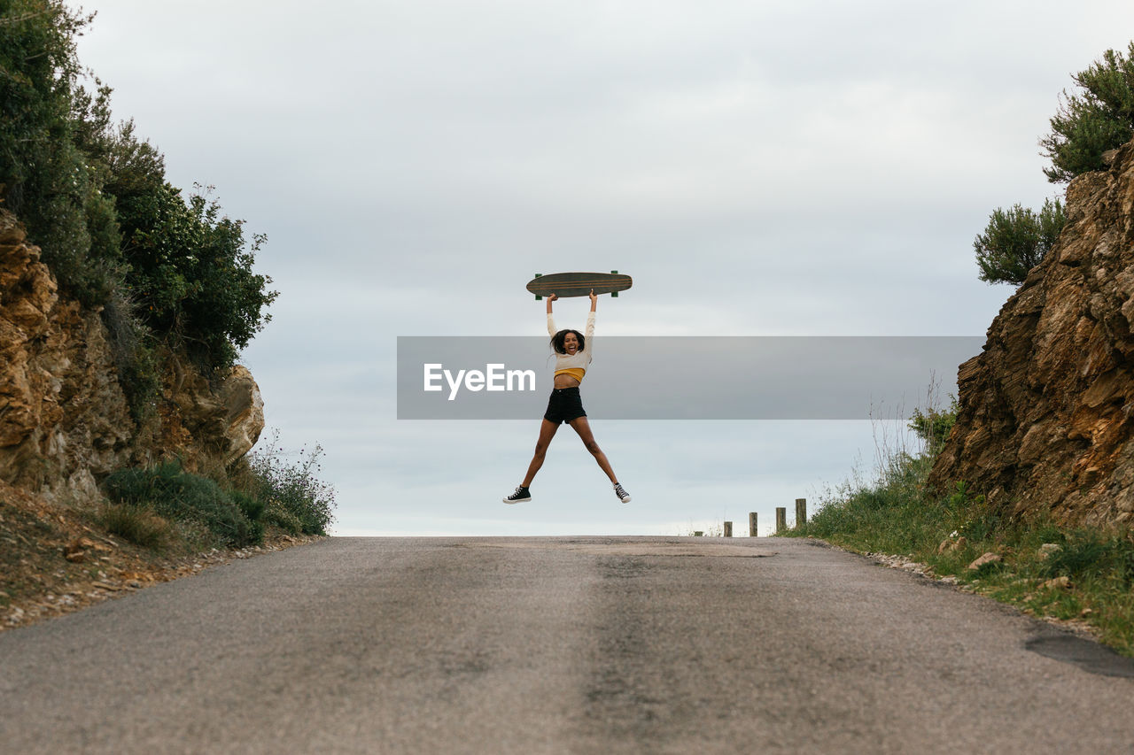 Full body of positive ethnic female jumping high and raising hands with longboard on asphalt road