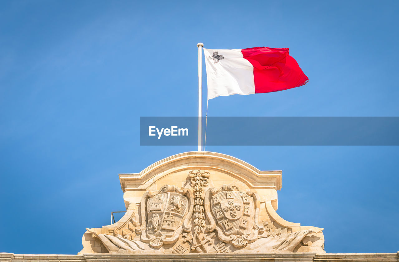 Low angle view of maltese flag on building against blue sky