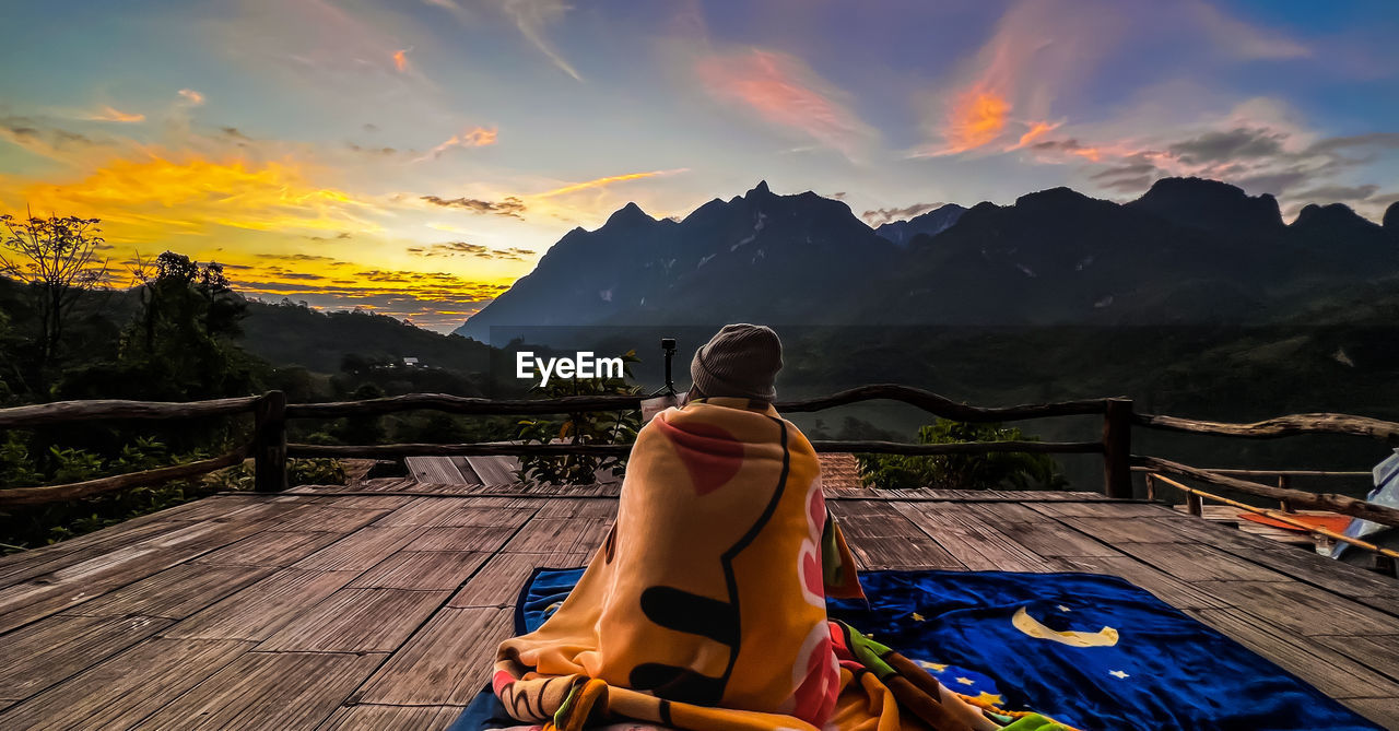 Rear view of woman sitting on boardwalk against sky during sunset