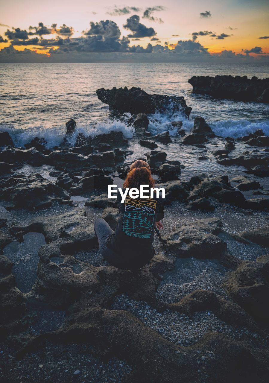rear view of man sitting on beach against sky during sunset