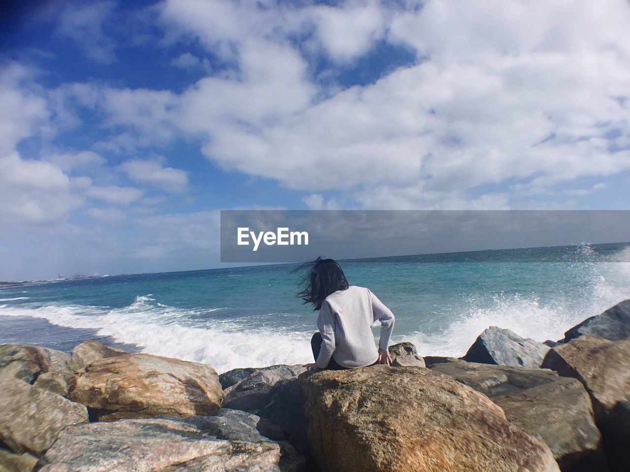 Rear view of woman sitting on rocks beach against sky