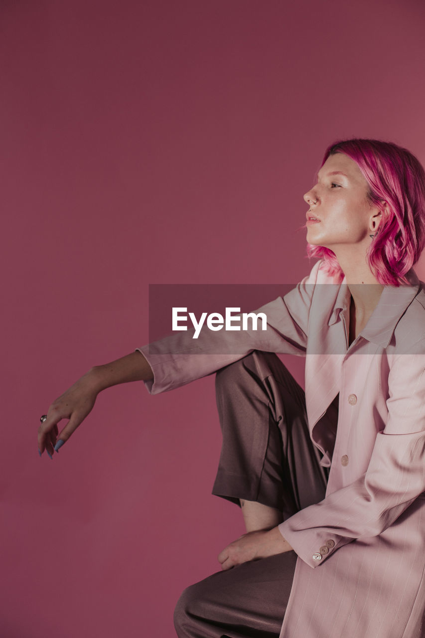 Young woman wearing coat while sitting against pink background