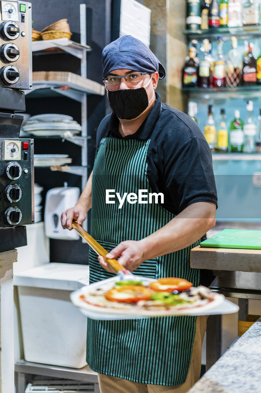 Portrait of male chef wearing protective face mask holding peel with freshly made pizza