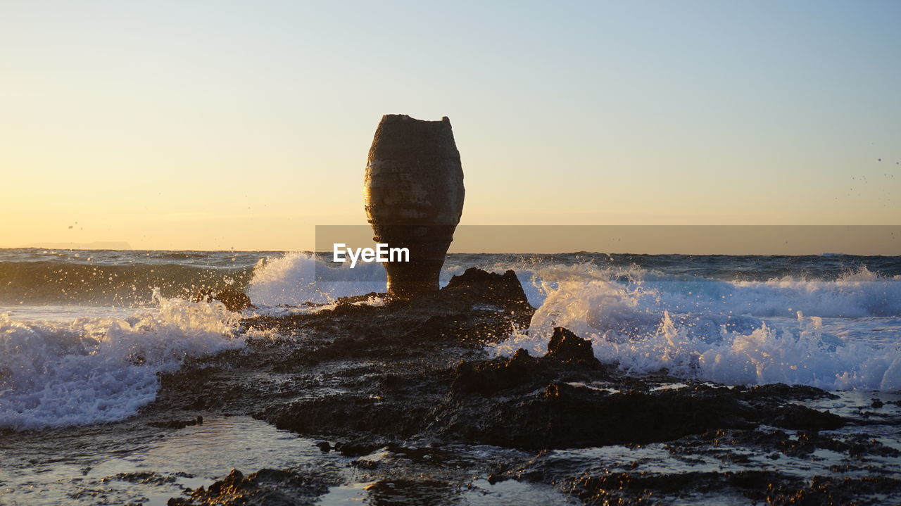 Sea waves splashing on rocks against sky during sunset