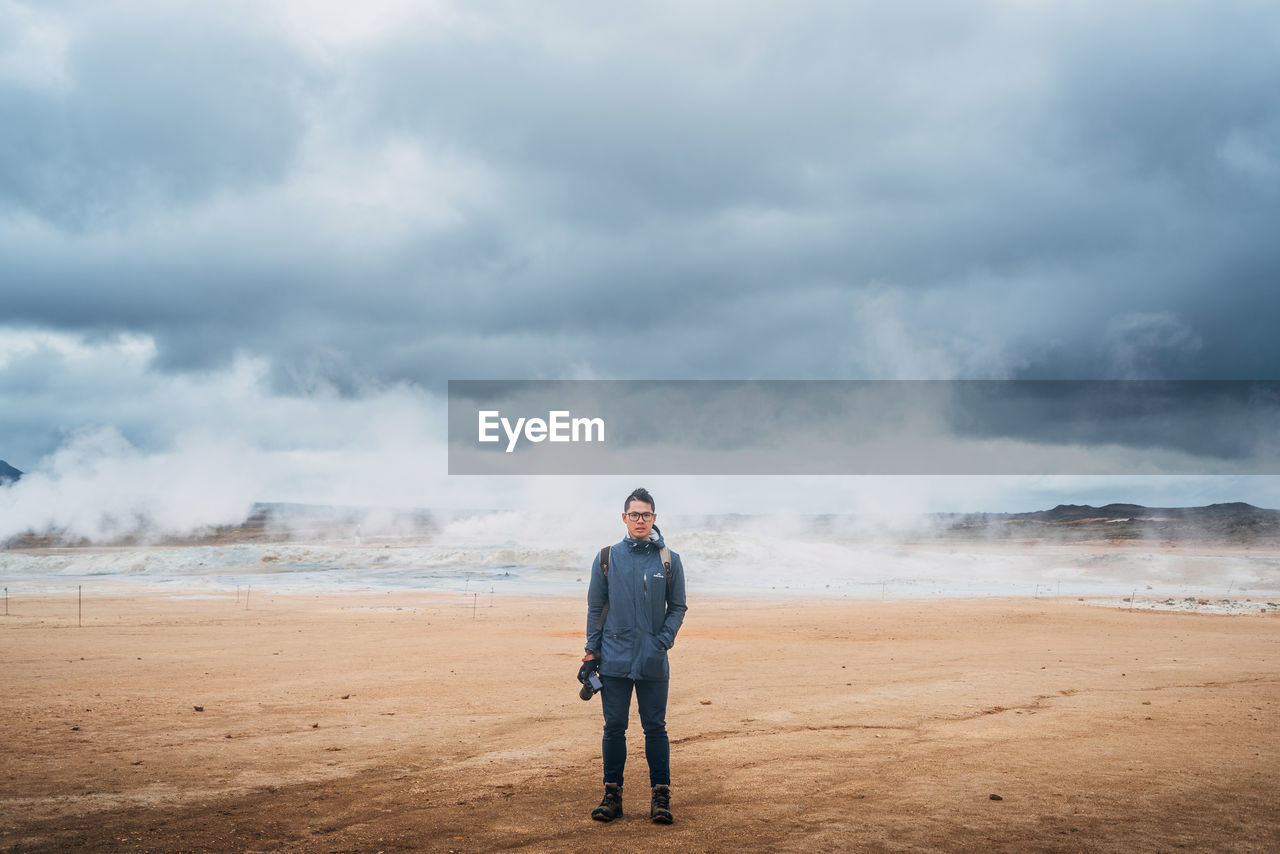 Full length of young man standing by hot springs on field against cloudy sky