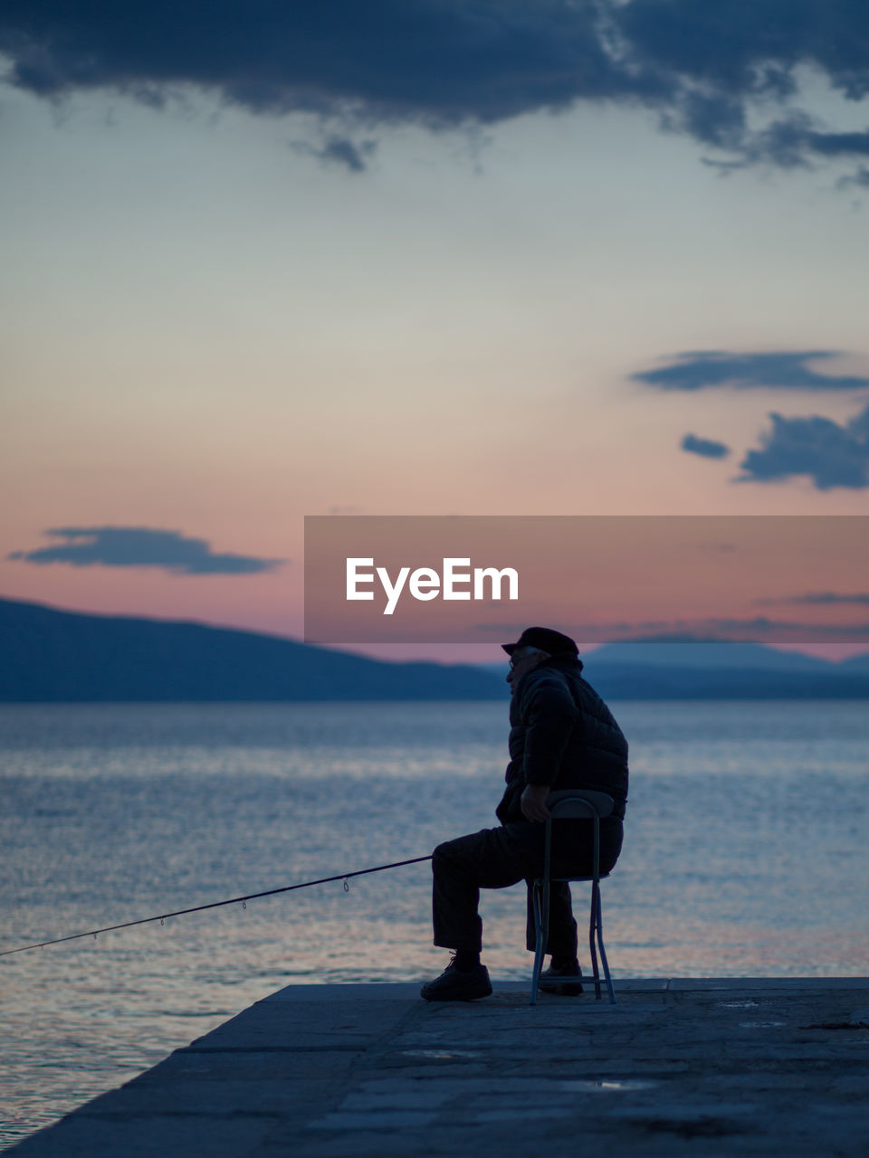 Man sitting on shore against sky during sunset