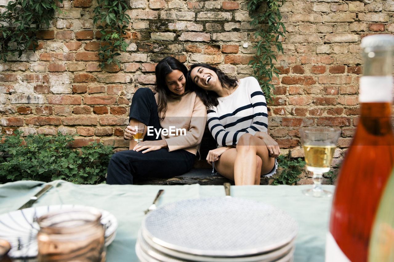 Happy female friends enjoying together while sitting on bench against wall at dinner party