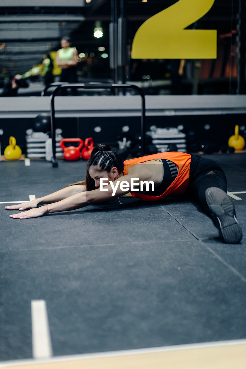 Full body young sporty female in fitness outfit performing stretching exercise on the floor while warming up during training in gym