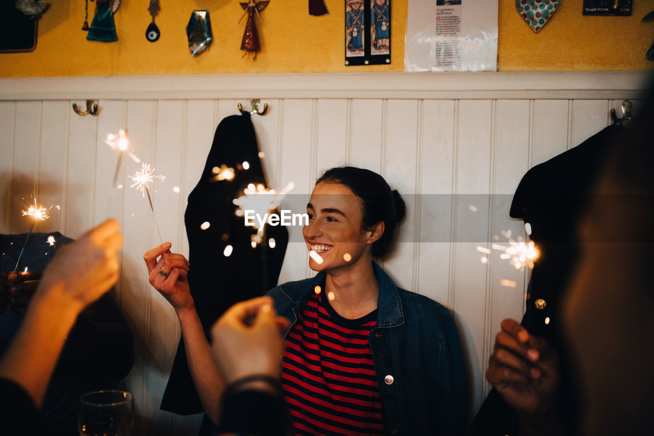 Smiling young woman holding burning sparkler while enjoying with multi-ethnic friends in restaurant during dinner party