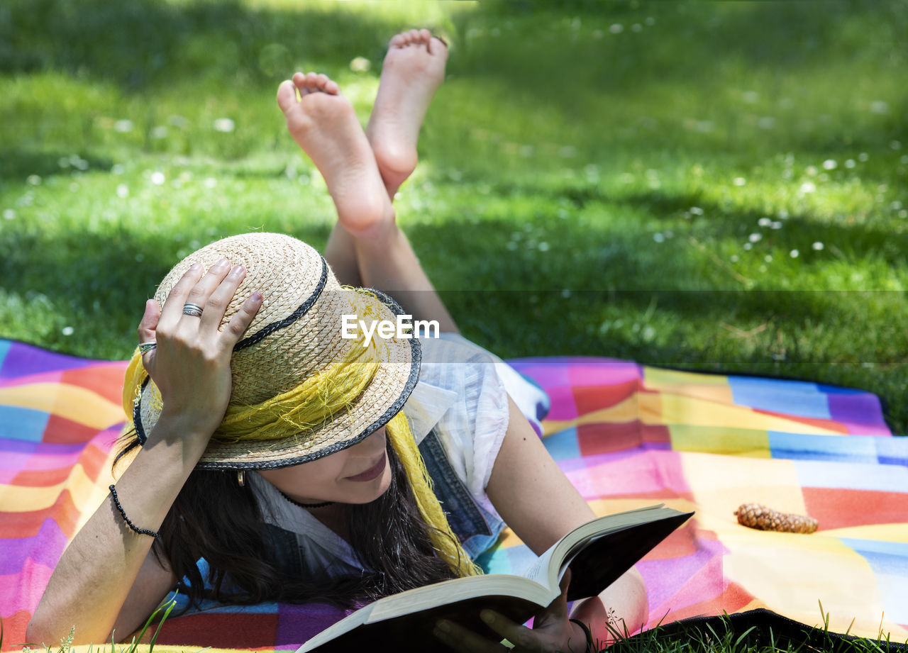 Full length of woman reading book while relaxing on picnic blanket at field