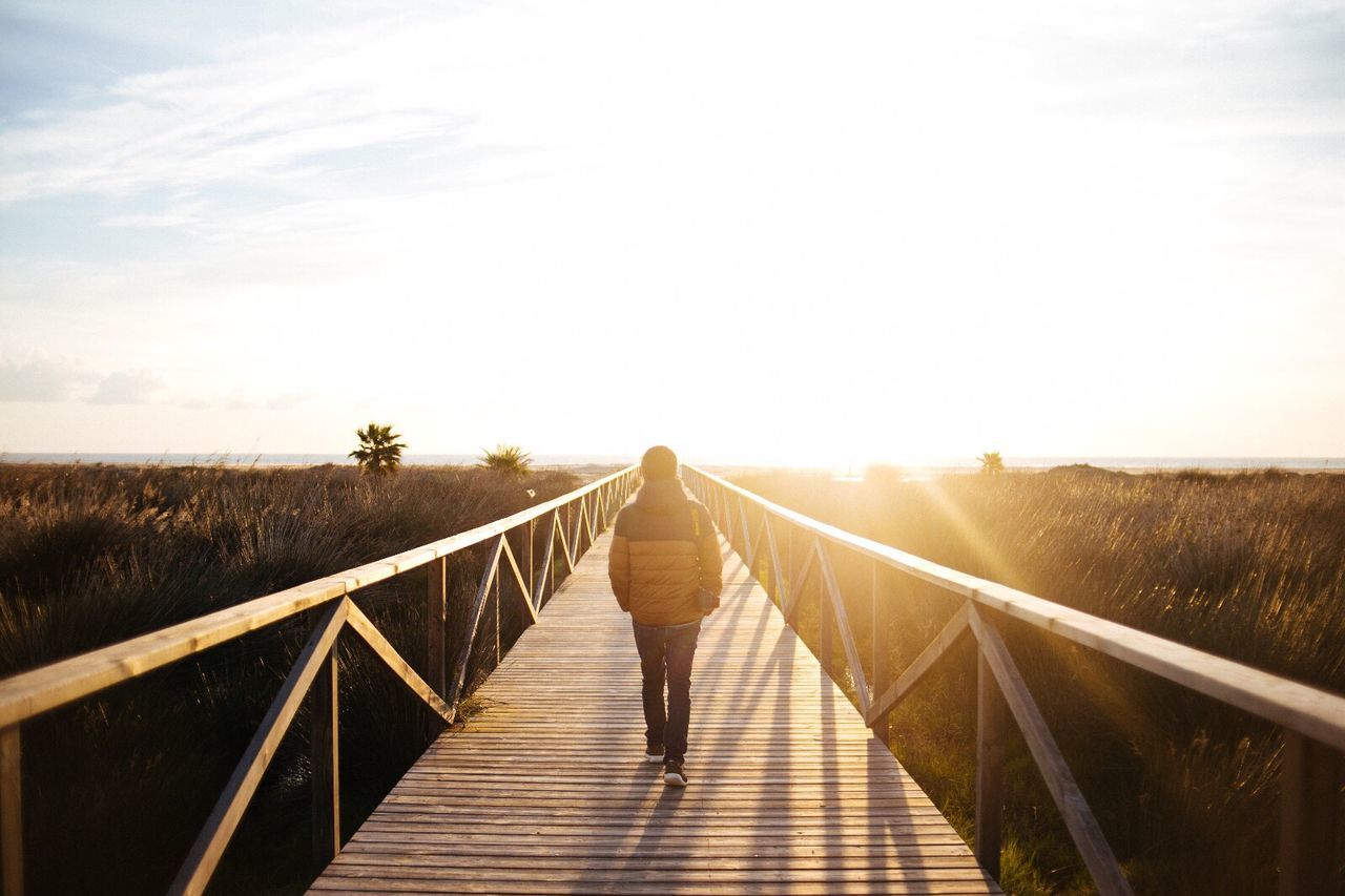 REAR VIEW OF WOMAN WALKING ON FOOTBRIDGE ALONG SKY