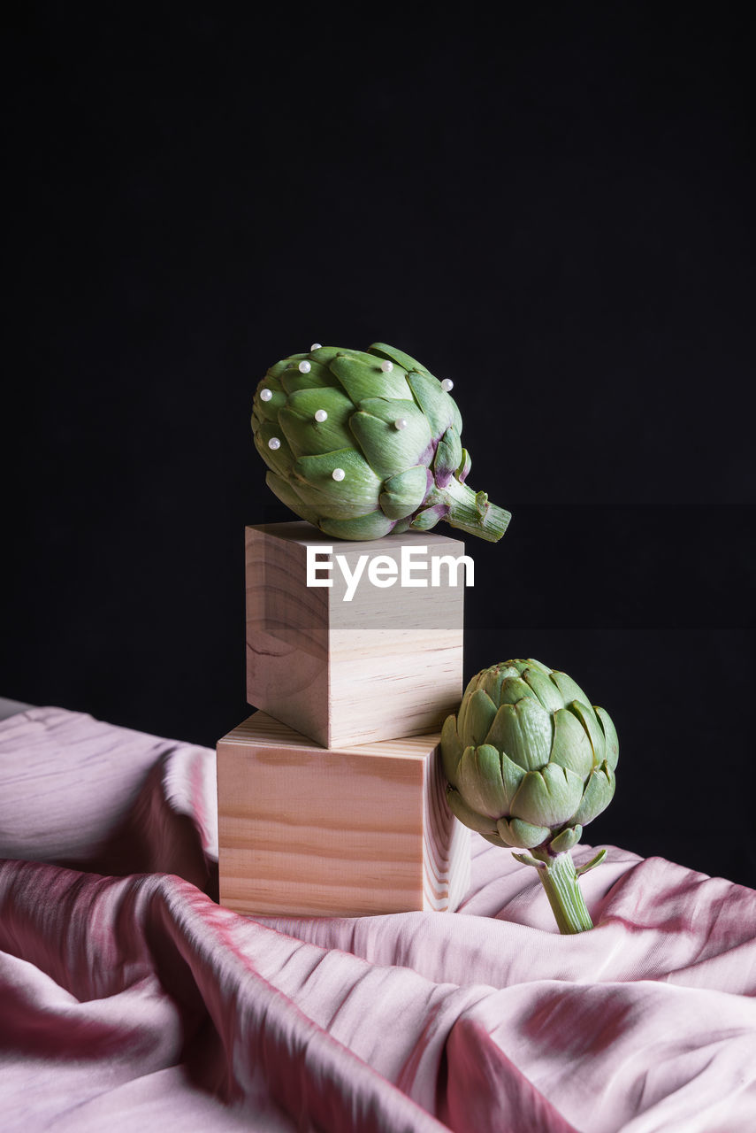 Ripe artichokes placed on wooden cube on table with silk fabric against black background