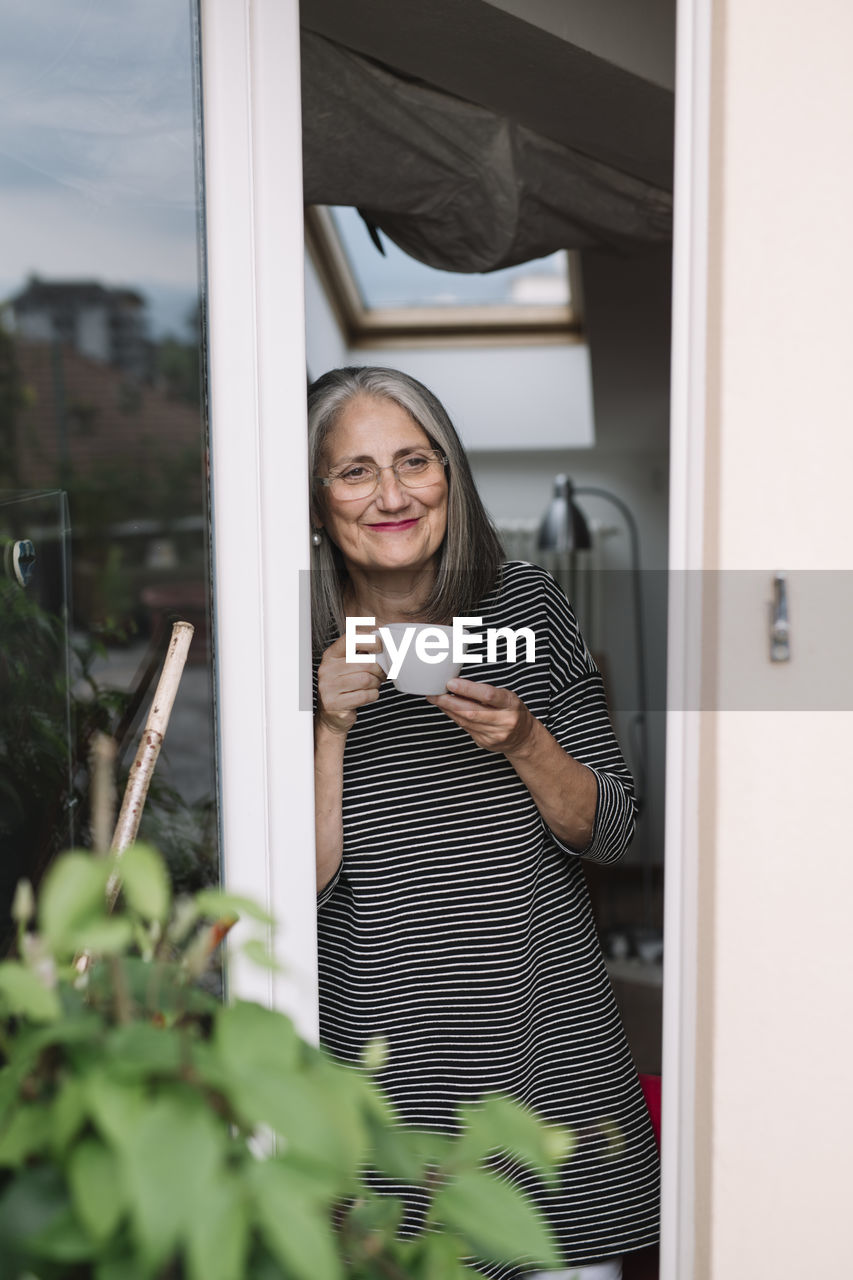 Portrait of happy senior woman leaning at opened balcony door with sup of coffee looking at distance