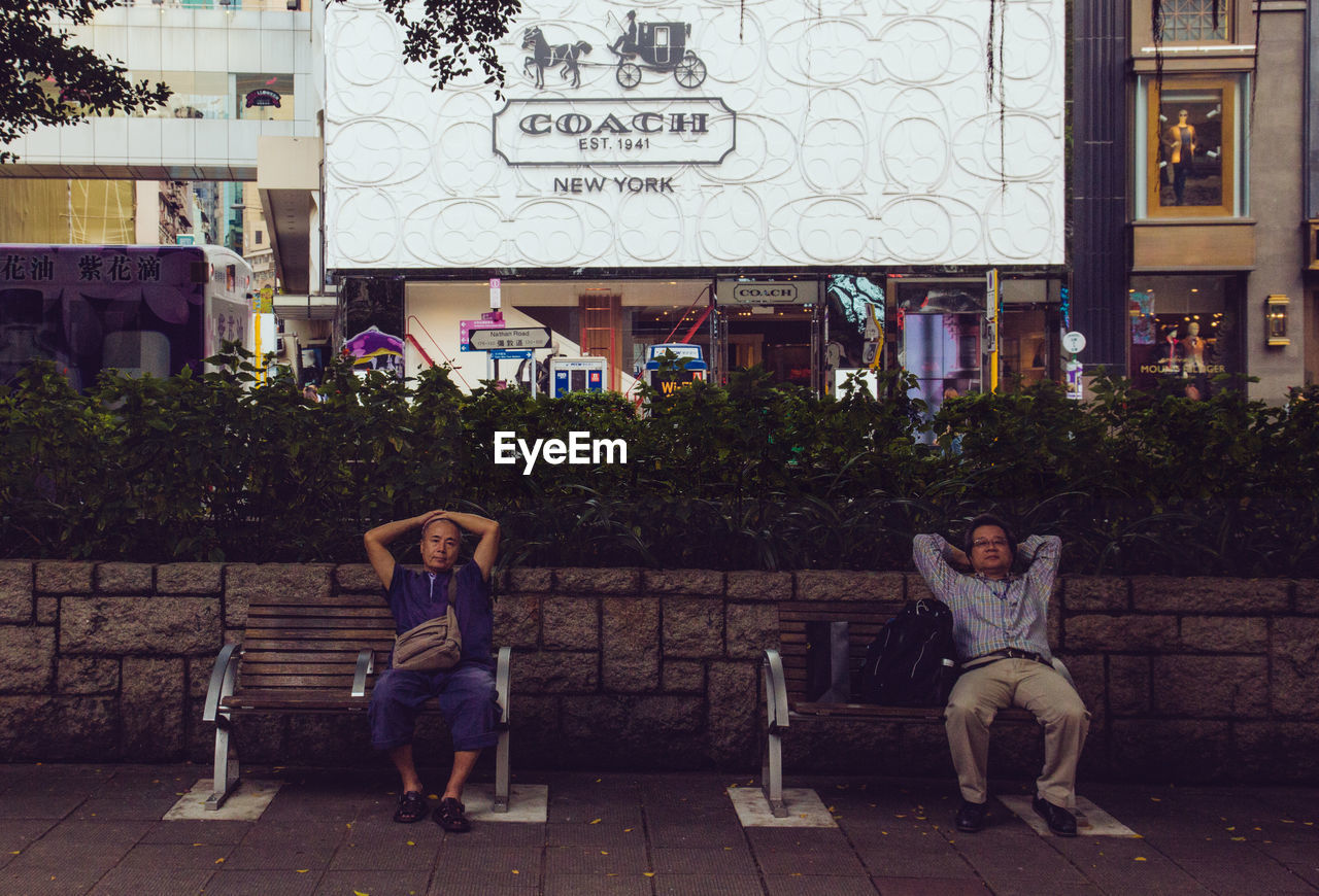 PEOPLE SITTING ON BENCH AGAINST BUILDING IN CITY