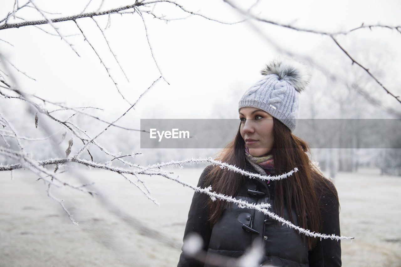 PORTRAIT OF YOUNG WOMAN STANDING BY BARE TREE